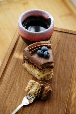 Piece of Kiev cake with chocolate icing and blueberries and cup of coffee on a wooden board. A bite...