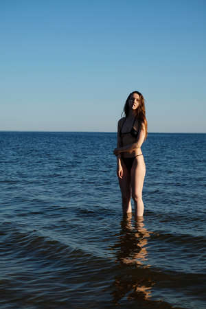 A girl in a black bikini with a cigarette in her mouth stands in the sea. summer landscape. fashion photoの写真素材