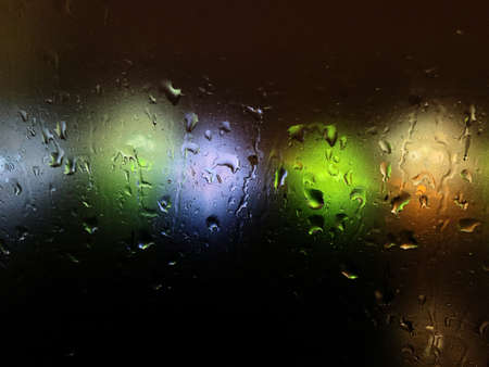 Colored blurry lights on a dark background through the drops on the windowの写真素材