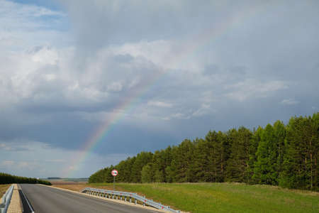 Landscape empty track road along green fields. Rainbow in the after-rain sky