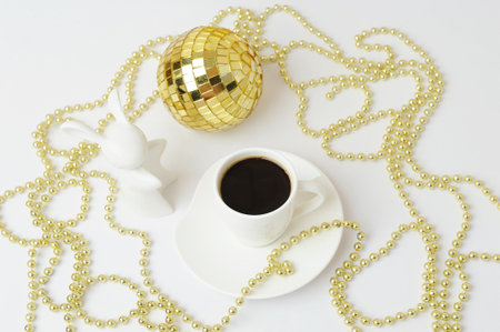 Cup of black espresso and white ceramic rabbit. Symbol of the year. Gold Christmas beads and disco ball. New year concept. white backgroundの写真素材