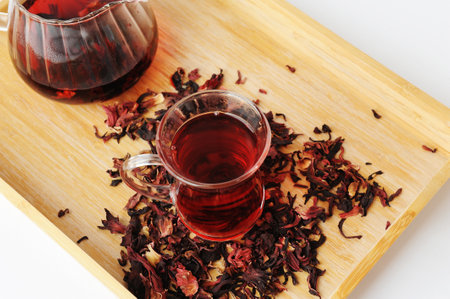 Pomegranate flowers tea in glass cup close up. teapot in the background. Bamboo tray with scattered petals. Beautiful ruby infusion. Flower drink, healthy food conceptの写真素材