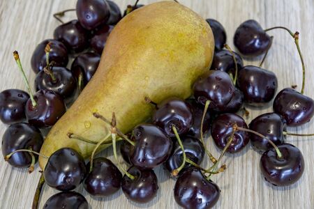 Still life. Big pear surrounded by a scattering of sweet cherry on a light wood backgroundの写真素材