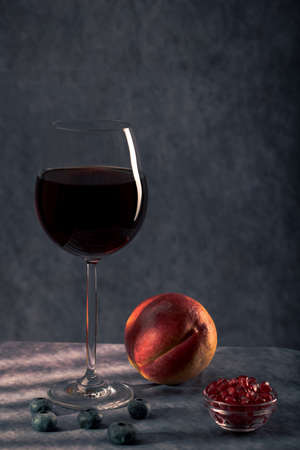 Still life. large glass with red wine, peach, pomegranate grains and blue berries on a dark background with creative lightingの写真素材