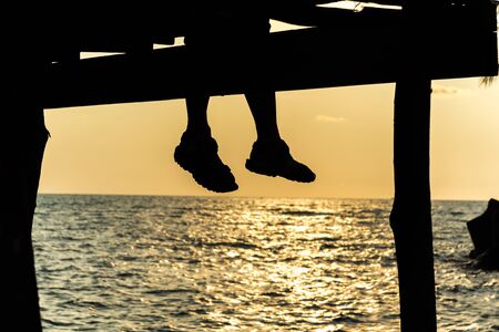 silhouette of feet of couple sitting on the pier at sunset beach,lonely.の写真素材