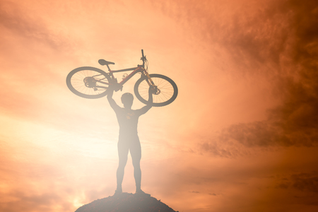 Silhouette the man stand in action lifting bicycle above his head on the Mountain with orange skyの写真素材