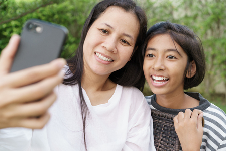 Asian mother and daughter selfie by smartphone together in the garden at home. A girl and women are smile when taking picture on garden backgroundの写真素材