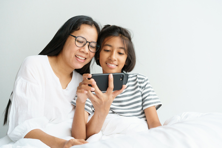 Asian mother and daughter playing game with phone together on the bed. Women and girl so happy and fun in bedroom.の写真素材