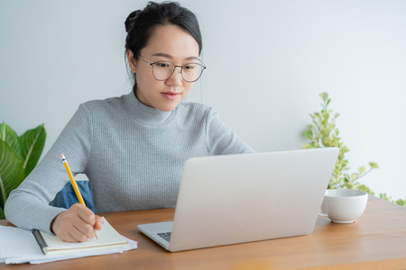 Asian woman wearing glasses is using laptop at home office.Portrait young cute student working on smart technology gadgetの写真素材