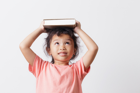 Asian little girl holding a book on head and eyes looking top on white background head. On face a cute girl so happy and smile.の写真素材