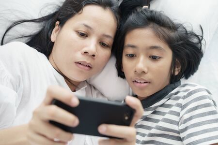 Asian mother and daughter playing game with phone together on the bed. Women and girl so happy and fun in bedroom.の写真素材