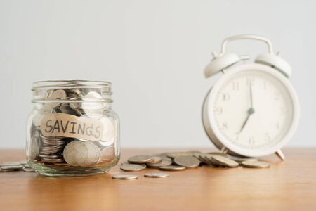 A coin in a glass bottle, a pile of coins and a white retro alarm clock on a brown wooden table. Investment business, retirement, finance and saving money for future concept.の写真素材