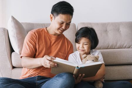 Asian father and daughter reading book while sitting in the living room at her home.の写真素材