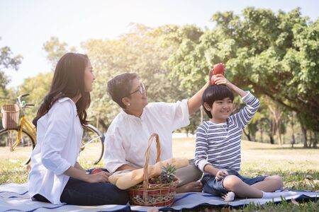 Asian family with son sitting hold apple above his head in the public park. Concept of lifestyle at happy in family holidayの写真素材