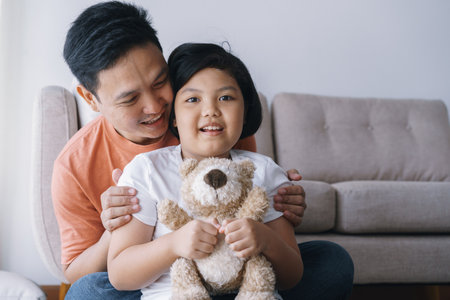 Happy Asian father and daughter playing together in the living room at home. The cute little girl hug the doll and smile happily with his father.の写真素材