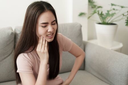 Asian woman have toothache pain while sitting on the sofa at home, Asia of girl on the face expression. Concept toothache and dental medicineの写真素材