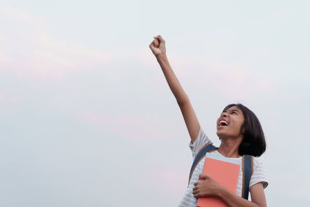 Happy Asian schoolgirl success and excited on the face while holding a book with hand raised up to blue sky background  in the summer at morningの写真素材