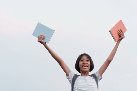 Happy Asian girl smile on the face while holding a book and hand raised up to blue sky backgroundの写真素材