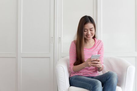 Beautiful young of Asian girl using a smartphone while sitting on the sofa in the living room at home, Asia women happy smile while chatting on mobile phoneの写真素材