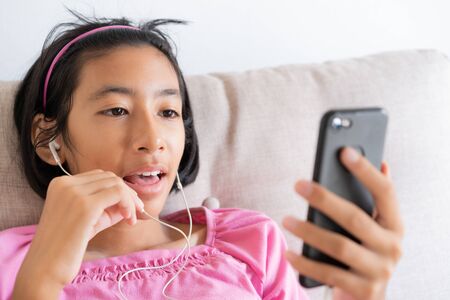 Asian girl video call on the smartphone while sitting on the sofa in the living room at home, Happy Asia child smile on the face and chat with earphone past internet. Lifestyle and relaxing conceptの写真素材