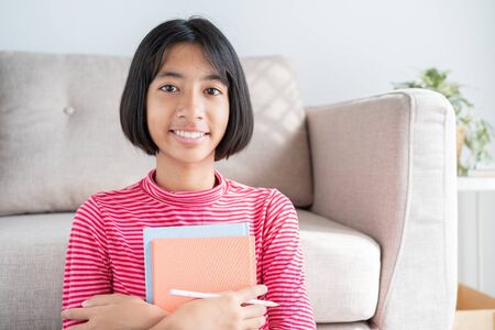 Happy Asian girl smile on the face while sitting in the living room at home morning, Portrait of Asia child hold book and happiness expression  confident. Concept learning at homeの写真素材