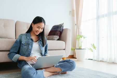 Asian girl wearing jeans using a laptop while sitting on the floor at home. Freelance girls are video conferencing with colleagues on social media. concepts work from home and new normalの写真素材
