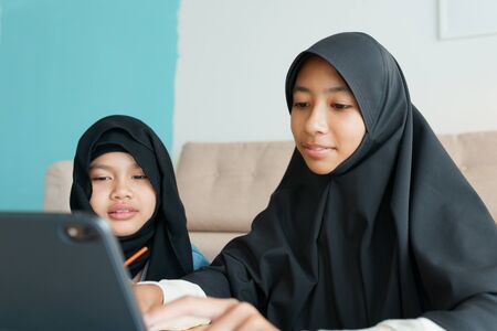 Two Muslim girl is studying online via the internet on tablet in living room at home, Asian elementary school children watching computer tablet. Concept of education at homeの写真素材