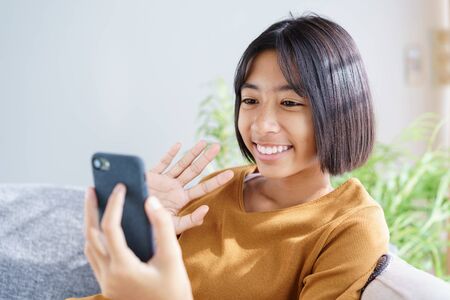 Happy Asian little girl video call on the smartphone and waving in greeting while sitting in the living room at home. Concept of relaxation and lifestyle at homeの写真素材