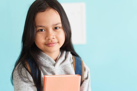 Happy Asian little girl smile on the face, Asia child hold book and backpack while standing in interior home. Portrait of a kid looking to camera. Concept backpack to schoolの写真素材