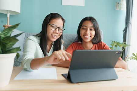 Asian family with a daughter do homework by using tablet with mother help. Happy smile Asia kid while sitting in the living room at home morning. Concept of online learning at homeの写真素材
