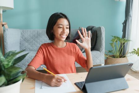 Happy Asian girl is studying online via the internet on tablet waving in greeting while sitting in living room at home morning. Asia child watch and write homework. Concept online learning at homeの写真素材