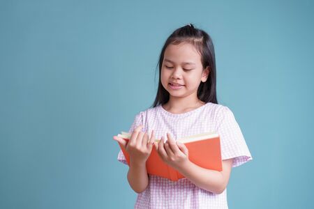happy Asian little girl standing reading the book on blue background isolatedの写真素材