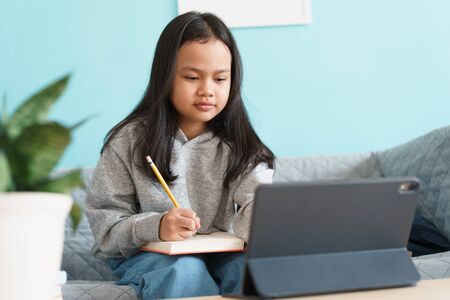 Asian little girl is studying online via the internet on tablet while sitting on sofa in living room at home. Asia children watch and write homework or practice. Concept of online learning at homeの写真素材
