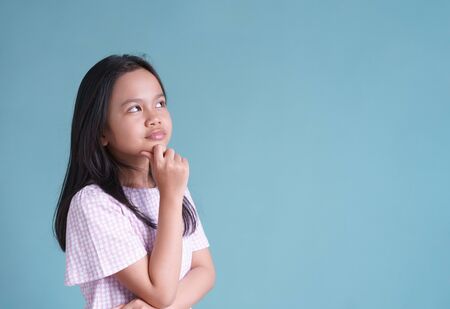 Asian little girl standing thinking and put hand under the chin her on blue background isolatedの写真素材