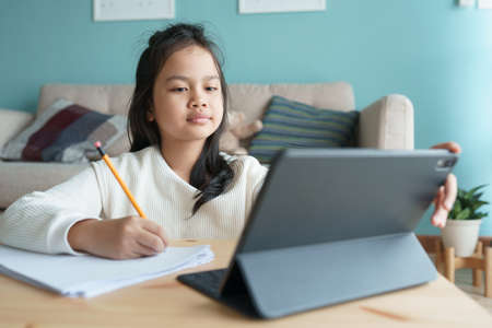 Education. Asian little girl is learning with online tutor via the internet on tablet digital at home morning. Asia children watch computer and doing homework. Concept of online learning at homeの写真素材