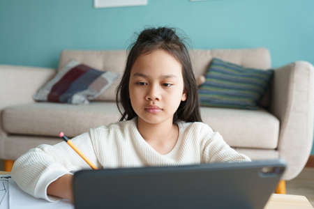 Education. Asian girl is learning with online tutor via the internet on tablet digital at home morning. Asia children watch computer and write homework. Concept of online learning at homeの写真素材