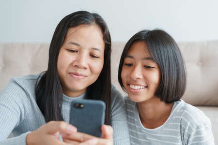 Family Asian mother and daughter watching entertainment online on smartphone together at home. Happy girl and mum while sitting in the living room togetherness. Close-up on face child and momの写真素材