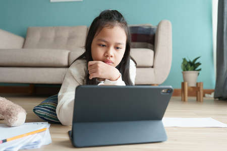Education. Asian girl is learning with online tutor via the internet on tablet digital in morning. Asia children doing homework with computer while lying on floor. Concept of online learning at homeの写真素材