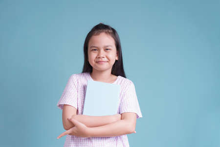 happy Asian little girl standing holding the book  on blue background isolated