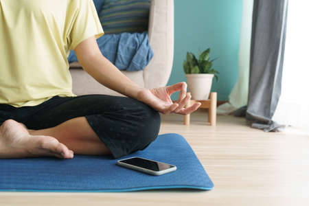 Hand of woman in lotus pose while sitting at mat yoga, Asian women practice yoga. With an online Trainer on mobile phone at home. Concept of relaxation and meditationの写真素材