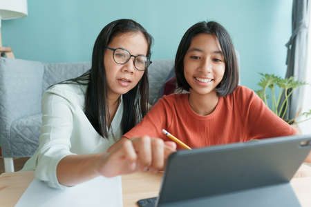 Asian family with a daughter do homework by using tablet with mother help. Happy smile Asia kid while sitting in the living room at home morning. Concept of online learning at homeの写真素材