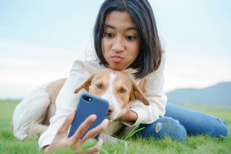 happy Asian girl Take a photo a dog and playing with smartphone On the green lawn together close-up at morningの写真素材