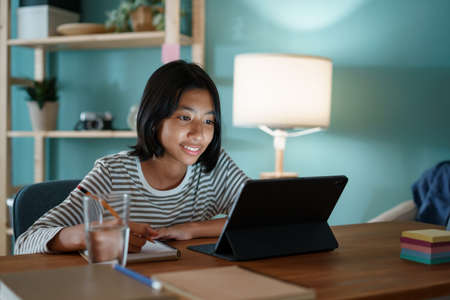 Homeschooling Asian girl doing homework and study online with tutor on tablet digital at home at nightの写真素材