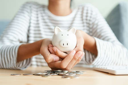 Hand of Asian young girl holding white piggy bank  with two hands on the coin stack at home morning, retirement, finance and saving money for future conceptの写真素材