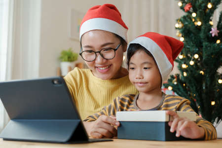 Christmas. Asian family with a mother and daughter giving gift box via video call together with tablet at homeの写真素材
