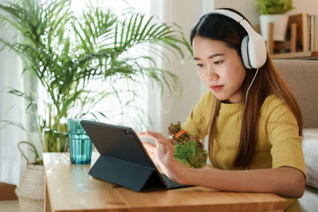 beautiful Asian young woman using a tablet and eat salad for lunch on table work at home. concept working from homeの写真素材