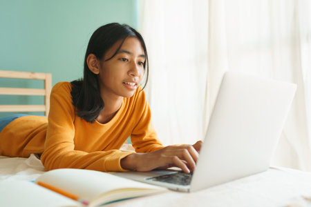 Asian woman learning online with a tutor via the internet with a laptop, Asia student girl video conference on a computer while lying on a bed in the morning, Concept online learning at homeの写真素材