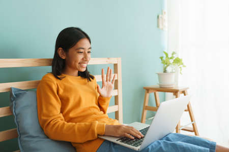 Happy Asian girl waving in greeting online tutor on a laptop computer in the interior home. Concept of online learning at homeの写真素材