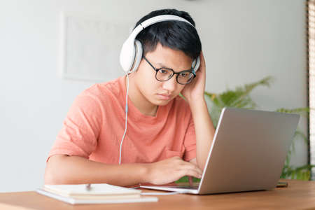 Asian student man learning online using a computer laptop with headphones while sitting workplace on a table at home in the morning. Concept online learning at homeの写真素材