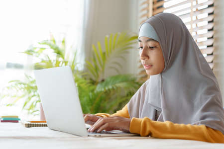 Side view of Muslim student girl wear hijab while sitting typing work on a computer laptop at home. Concept online learning at homeの写真素材